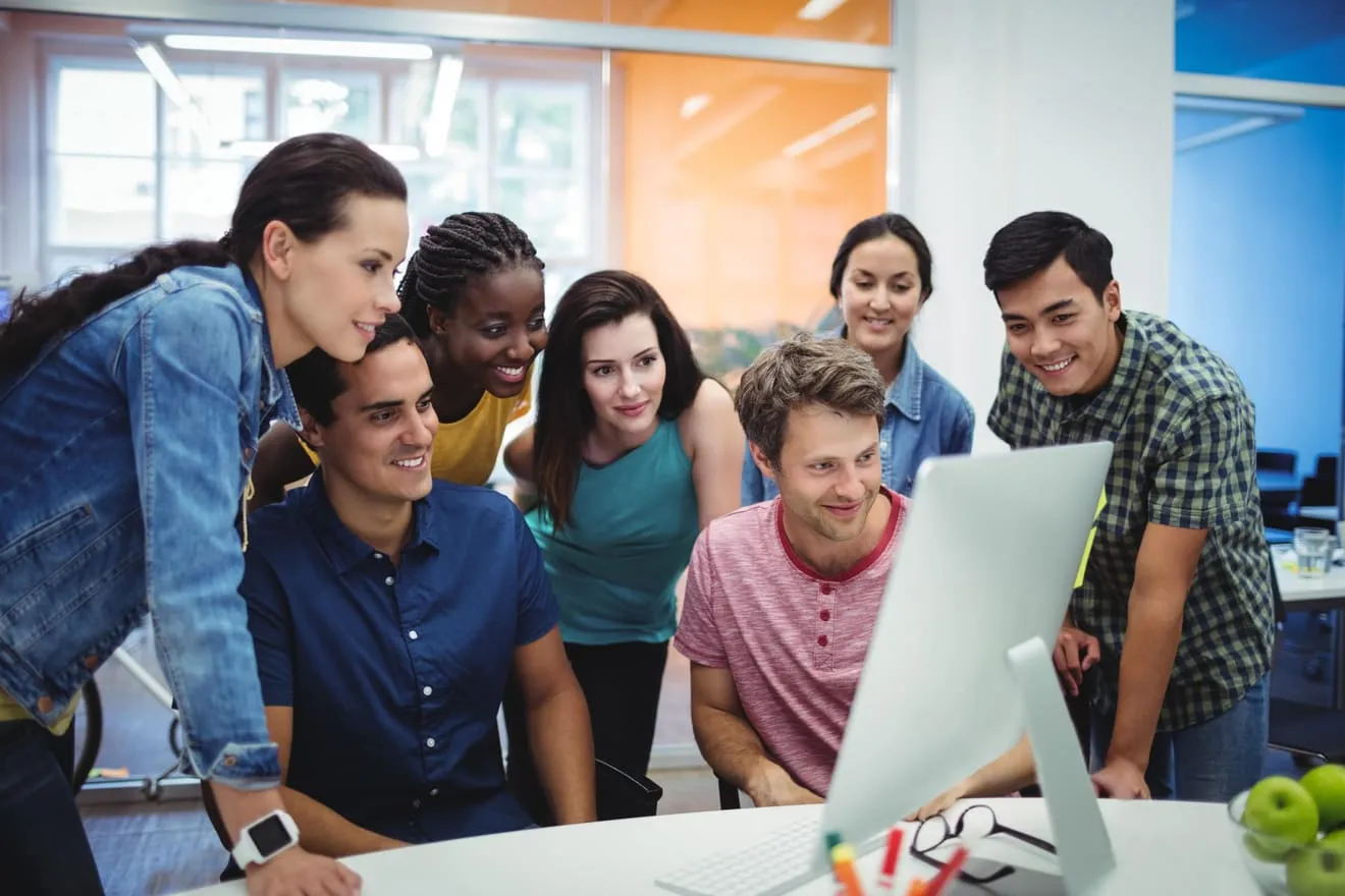 Grupo de siete jóvenes profesionales sonrientes, reunidos alrededor de la pantalla de un ordenador en una oficina.
